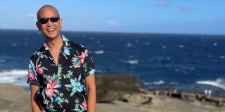 A man in a Hawaiian shirt standing on a rocky beach with the ocean in the background